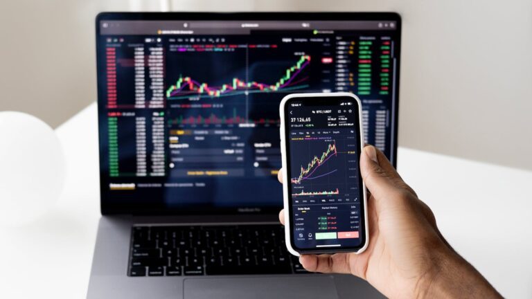 Eine Person hält ihr Smartphone in die Kamera, dahinter ein Laptop, auf beiden sind Aktienkurse zu sehen